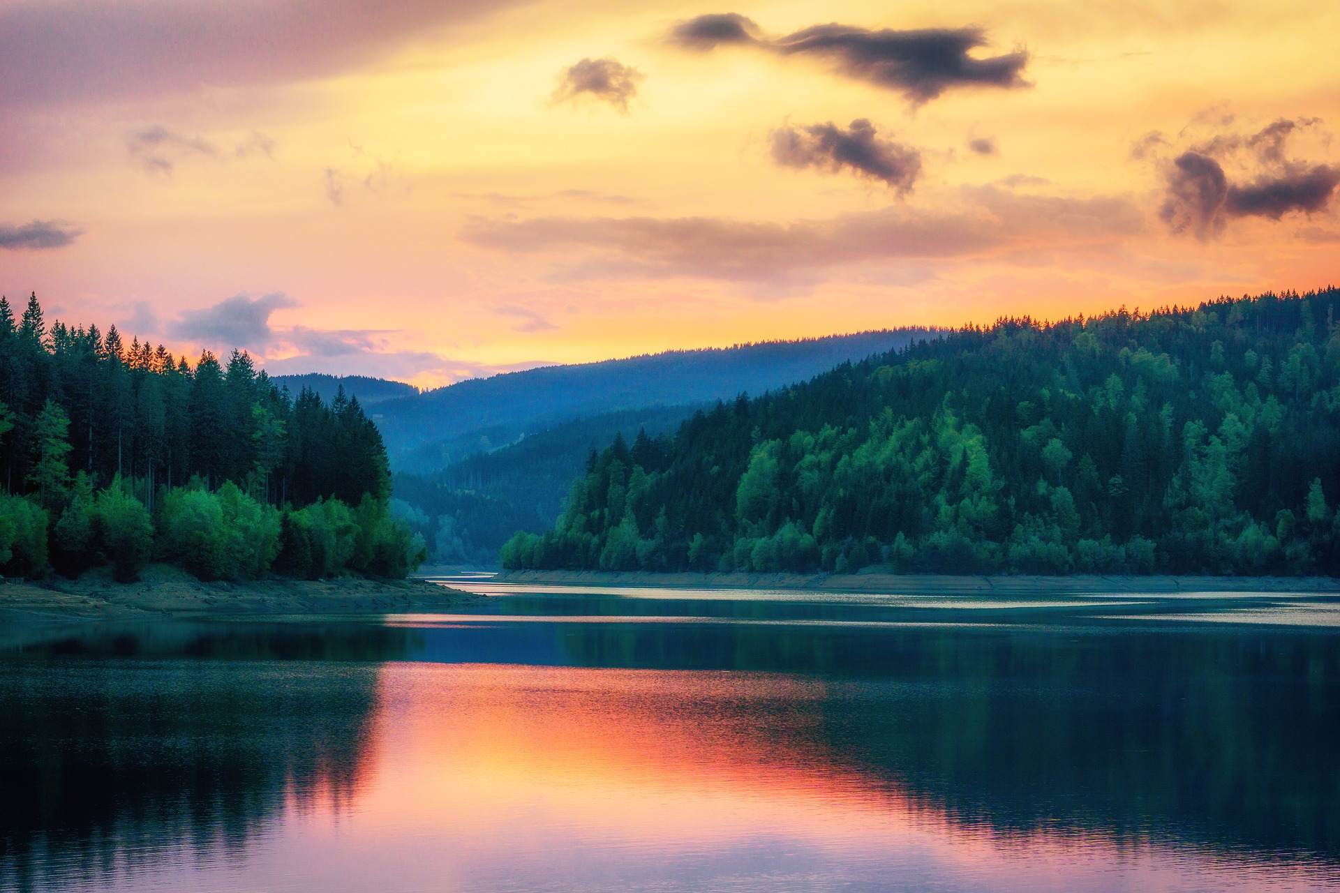 Ein Blick auf einen See bei Sonnenuntergang. Im Hintergrund sind Bäume und Hügel zu sehen